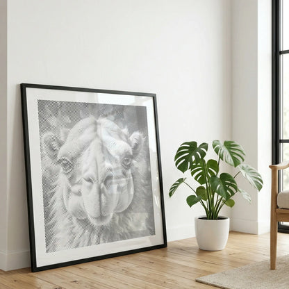A large black-framed black and white art print of a camel's face leaning against a white wall on a light wood floor, next to a potted green plant and a wooden chair. Camel | Dot Painting Kit